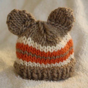 May include: A hand-knitted baby hat in shades of brown, orange, and white. The hat features two small ear-like protrusions at the top. The design includes horizontal stripes of orange and white, creating a cozy and playful look.