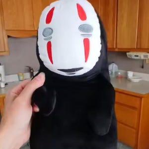 Puede incluir: Un peluche del personaje Sin Rostro de la pel&iacute;cula de animaci&oacute;n El Viaje de Chihiro. El juguete tiene una cara blanca con marcas rojas y formas de ojos grises. El cuerpo es negro y el juguete lo sostiene una persona.