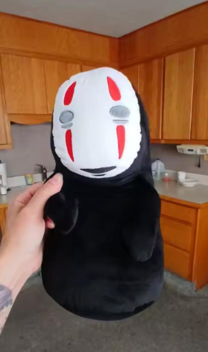Puede incluir: Un peluche del personaje Sin Rostro de la pel&iacute;cula de animaci&oacute;n El Viaje de Chihiro. El juguete tiene una cara blanca con marcas rojas y formas de ojos grises. El cuerpo es negro y el juguete lo sostiene una persona.