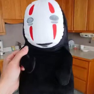 Puede incluir: Un peluche del personaje Sin Rostro de la pel&iacute;cula de animaci&oacute;n El Viaje de Chihiro. El juguete tiene una cara blanca con marcas rojas y formas de ojos grises. El cuerpo es negro y el juguete lo sostiene una persona.