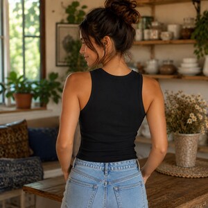 Puede incluir: Un top sin mangas negro acanalado y vaqueros de mezclilla azul claro. La mujer est&aacute; de espaldas a la c&aacute;mara, con el pelo recogido en un mo&ntilde;o. El fondo incluye una mesa de madera, plantas y una ventana.