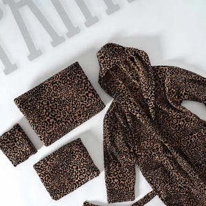 Op de afbeelding: Een set badtextiel met een bruin en zwart luipaardpatroon. De collectie bestaat uit een badjas met capuchon, lange mouwen en een ceintuur, een grote rechthoekige badhanddoek en twee kleinere vierkante washandjes of handdoeken.