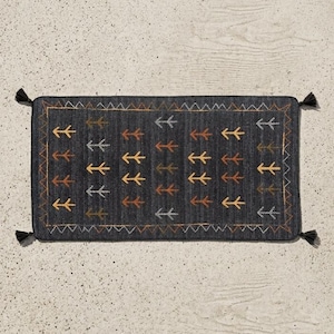 Op de afbeelding: Een rechthoekig, donkergrijs tapijt met een herhalend pijlenpatroon in tinten geel, bruin en grijs. Het tapijt heeft een zigzagrand en kwastjes op elke hoek. Het tapijt is ongeveer 91 cm lang en 61 cm breed.