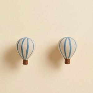 Puede incluir: Dos pomos decorativos de globo aerostático. Cada pomo presenta un globo blanco con rayas verticales azules y una base marrón. Los pomos están diseñados para montarse en una pared o mueble, añadiendo un toque caprichoso a cualquier habitación.