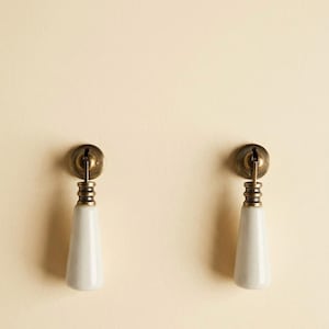 Può includere: Due pomelli per mobili con manico in ceramica bianca a forma di cono e ferramenta in ottone anticato. I pomelli sono montati su una superficie color crema, che presenta un design classico per mobili o decorazioni.