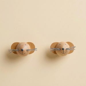 Könnte beinhalten: Zwei mausförmige Wandhaken aus Holz. Jeder Haken hat einen runden Kopf mit einer aufgemalten schwarzen Nase und einem lächelnden Mund. Die Ohren sind abgerundet und die Schnurrhaare bestehen aus dünnem, grauem Draht. Die Haken sind aus hellem Holz gefertigt.