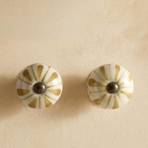 May include: Two ceramic cabinet knobs. Each knob is round with a white base and a pattern of gold-colored petal shapes radiating from a dark brown center. The knobs are set against a light beige background.