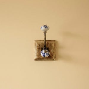 Puede incluir: Un gancho de pared decorativo con una base de madera cuadrada y un brazo metálico. Cuenta con dos pomos de cerámica blanca con diseños de hojas azules. Este gancho está diseñado para colgar objetos y añade un toque rústico a cualquier estancia.