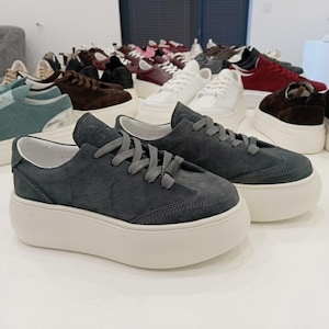 May include: A pair of dark gray suede sneakers with thick, off-white soles. The shoes have gray laces and a low-profile design. Other sneakers in various colors, including white, brown, and burgundy, are visible in the background.