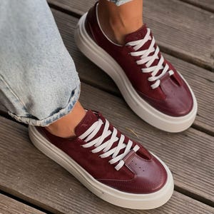 Può includere: Un paio di sneakers basse color bordeaux intenso con tomaia in camoscio e pelle, lacci bianchi e una spessa suola platform in gomma bianca. Il design presenta una punta rinforzata con piccoli fori di ventilazione e una linguetta con logo.