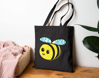 May include: Black tote bag featuring a cartoon bee with yellow and black stripes, a smiling face, and the text "BEE YOU" in blue. The bag has black handles and is made of a durable fabric.