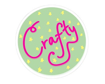 May include: A round sticker with the word "crafty" in bright pink cursive lettering. The word is centered on a light green background scattered with small yellow stars. The sticker has a white border.