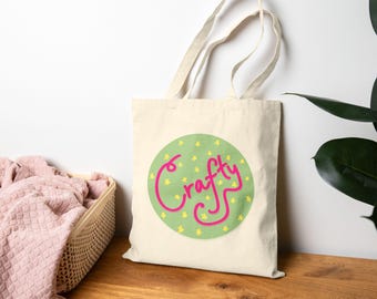 May include: Cream-colored tote bag with a green circle featuring yellow stars and the pink word "Crafty". The bag has long handles and is made of a sturdy fabric, suitable for carrying various items. The design is simple and eye-catching.