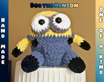 May include: Handmade crochet Minion plush toy in yellow, blue, and black. The plush features large white and gray eyes, and the text "Bob the Minion" and "One of a Kind" are visible.