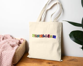 May include: Cream-colored tote bag with the word "crochete" in colorful, cut-out style letters. The bag has a simple design with two handles and is made of a sturdy fabric. The bag is sitting on a wooden surface.