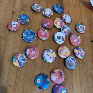 May include: A selection of circular button badges with diverse cartoon character illustrations. The badges display a range of colours, such as red, blue, yellow, and white. The designs include faces, abstract patterns, and other graphic elements.