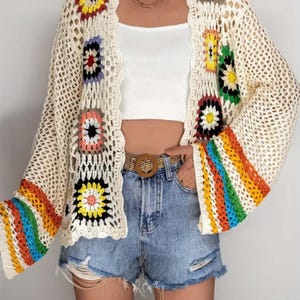 Könnte beinhalten: Cremefarbene Häkeljacke mit bunten quadratischen Mustern und Regenbogenstreifen an den Ärmeln. Der offene Pullover hat ein lockeres, luftiges Gewebe und einen gewellten Rand. Die Ärmel sind ausgestellt, und die Jacke wird mit Jeansshorts getragen.