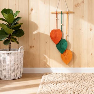 May include: A macrame wall hanging with three leaf shapes in orange and green, suspended from a wooden dowel. The leaves are attached to cords with wooden beads. A potted plant in a white basket and a beige rug are in the foreground.
