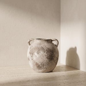 Peut inclure: Un vase en céramique rond doté de deux petites anses latérales. Le récipient présente une finition texturée et patinée dans des tons beige, crème et brun sourd, créant un aspect tacheté sur tout son corps sphérique et son ouverture.