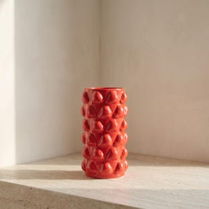 Peut inclure: Un vase cylindrique en céramique d'un orange vif, présentant un motif répétitif de bosses triangulaires arrondies et en relief sur toute sa surface extérieure, créant un aspect texturé et tactile avec une finition brillante.