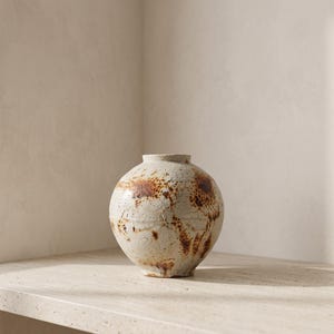 May include: A spherical ceramic vase with a textured, off-white matte finish. The surface features irregular, dark brown rust-coloured spots and patches distributed across the rounded body, with a small, circular opening at the top centre.