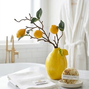 Peut inclure: Un vase jaune en forme de citron contient une branche avec des citrons et des feuilles vertes. Une serviette blanche pliée avec une étiquette et un pain de savon sur une petite soucoupe sont sur le comptoir. Un peignoir blanc est suspendu en arrière-plan.