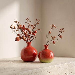 Könnte beinhalten: Zwei granatapfelförmige Keramikvasen in glänzendem Rot mit gesprenkelten Details. Die größere Vase enthält ein dichtes Bündel dünner Zweige mit kleinen roten Beeren und Blättern, während die kleinere Vase einen einzelnen geschwungenen Zweig hält.