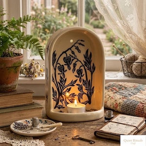 Peut inclure: Un photophore en céramique crème avec un motif floral noir. Une bougie chauffe-plat allumée à l'intérieur diffuse une lumière chaude. Le photophore est posé sur une surface en bois avec une petite assiette, des livres et une fougère en arrière-plan. Le texte "Quiet Rituals Craft" est visible.
