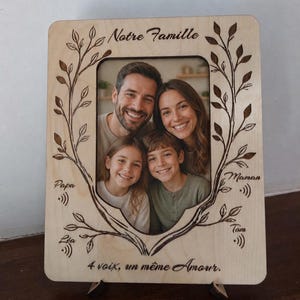 Peut inclure: Cadre photo familial en bois clair, de forme rectangulaire. Il présente une photo de famille entourée d'un motif de feuilles. Le texte comprend "Notre Famille" et "4 voix, un même Amour."