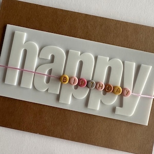 Op de afbeelding: Een rechthoekige wenskaart met een bruine basis en een wit verhoogd plaatje met het woord happy in grote letters. Een dun roze touwtje loopt over het midden en houdt kleine ronde kralen vast die het woord birthday spellen.