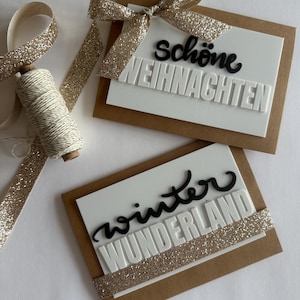 Può includere: Due biglietti d'auguri rettangolari con basi in cartoncino marrone e sovrapposizioni rettangolari bianche. Un biglietto presenta un fiocco glitterato dorato e il testo in bianco e nero "schöne WEIHNACHTEN". L'altro biglietto presenta il testo in bianco e nero "winter WUNDERLAND" con una striscia glitterata dorata.