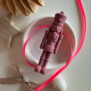 Puede incluir: Un adorno de cascanueces de color malva mate con un sombrero alto, un uniforme con botones y un pequeño lazo en la parte superior para colgar. La figura tiene una superficie texturizada y está colocada dentro de un pequeño plato redondo de cerámica blanca.