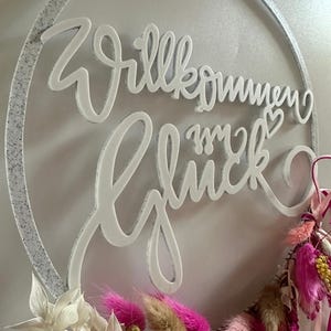 Könnte beinhalten: Ein weißer, gesprenkelter Ring mit dem weißen Schriftzug Willkommen im Glück. Der untere Teil des Rings ist mit einem Arrangement aus getrockneten Blumen in leuchtendem Pink, sanftem Beige und Gold verziert, ergänzt durch rosa Bänder.