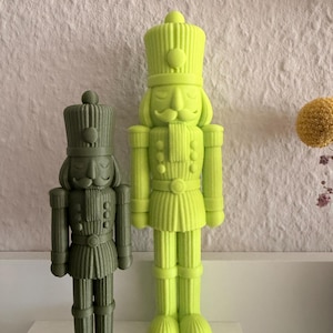 Pode incluir: Duas figuras de quebra-nozes lado a lado. A menor é verde-oliva escuro e a mais alta é amarelo neon brilhante. Ambas apresentam uma textura canelada, um chapéu alto, bigode e uma jaqueta estilo uniforme com botões detalhados.