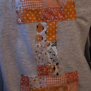 Può includere: Una felpa grigia con una grande lettera "I" patchwork applicata sul davanti. La "I" è composta da vari tessuti fantasia arancioni e bianchi, tra cui pois, quadretti e stampe floreali. Il design è un dettaglio unico, fatto a mano.