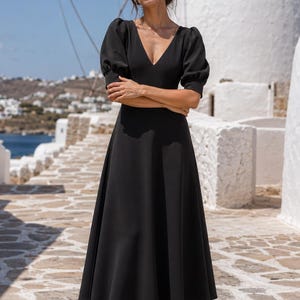 Könnte beinhalten: Ein einfarbig schwarzes Midikleid mit tiefem V-Ausschnitt, kurzen Puffärmeln mit eng anliegenden Bündchen und einem ausgestellten A-Linien-Rock, der unterhalb der Knie endet. Das Kleidungsstück besteht aus einem glatten, blickdichten Stoff.