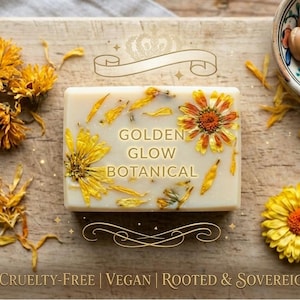 May include: A rectangular bar of soap with the text "GOLDEN GLOW BOTANICAL" and embedded yellow flowers and petals. The soap is on a wooden cutting board with dried flowers, a bowl of nuts, and a perfume bottle. The text "CRUELTY-FREE | VEGAN | ROOTED & SOVEREIGN" is at the bottom.