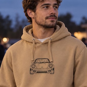 May include: A tan hooded sweatshirt featuring a black line art illustration of a sports car on the chest. The garment includes a front kangaroo pocket, long sleeves with ribbed cuffs, and a drawstring hood with metal eyelets.