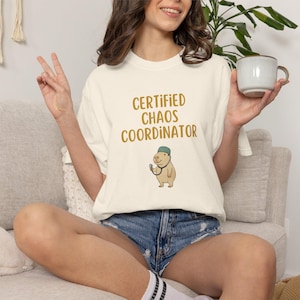May include: Cream-colored t-shirt with the text "CERTIFIED CHAOS COORDINATOR" in gold lettering. A cartoon capybara wearing a green hat and stethoscope is below the text. The person is wearing denim shorts.