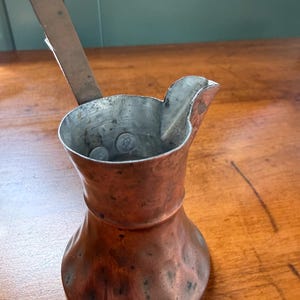 May include: A hammered copper coffee pot with a wide base, a narrow neck, and a flared rim featuring a pouring spout. The interior is lined with a silver-toned metal and includes a long, flat, metallic handle extending upward from the side.