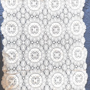 May include: A rectangular off-white lace runner featuring a repeating pattern of circular motifs with intricate floral-like centers. The lace has a scalloped edge and a delicate, open-work mesh structure throughout the entire textile piece.