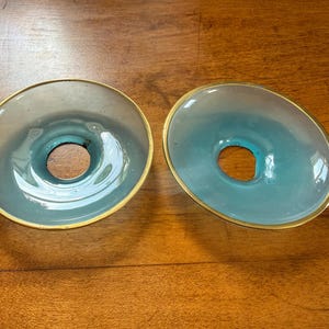 May include: Two circular glass lamp shades featuring a gradient of translucent white to light blue. Each shade has a smooth, curved surface and a central circular opening, finished with a thin, metallic gold-toned rim along the outer edge.