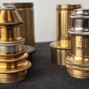 May include: Close-up of several vintage brass microscope objectives and components. The objectives have a gold finish with silver accents and intricate detailing. One objective is labeled "1/12" and "oil immersion". Other brass cylinders are in the background.