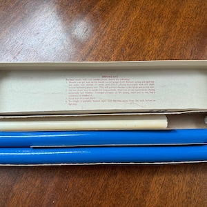May include: A rectangular cardboard box containing two bright blue cylindrical candle holders, a single off-white taper candle, and a small white ring. The inside lid features printed text titled IMPORTANT with instructions for candle maintenance and care.