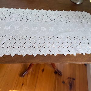 May include: A rectangular white table runner featuring intricate cutwork embroidery. The design includes a central row of interconnected circular patterns with small floral motifs, bordered by a scalloped edge with leaf-like cutouts.