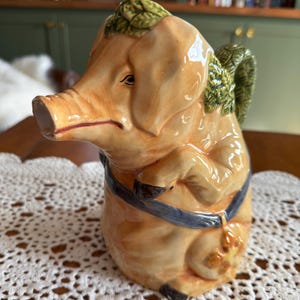 May include: A ceramic pitcher shaped like a pig with a glossy tan finish. The pig features a green leafy crown on its head, a green leafy handle, and a blue strap across its body with a small orange floral detail. The snout serves as the spout.