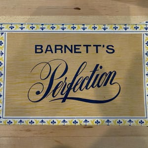 May include: A rectangular cardboard box with a light wood grain pattern. The text BARNETT'S is printed in blue, followed by the word Perfection in a large blue script font. The border features a repeating blue and yellow floral pattern.