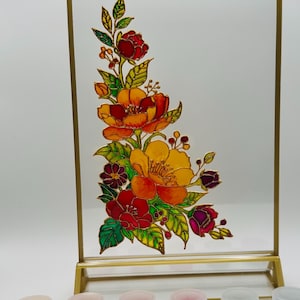 May include: A decorative glass panel with a gold frame, featuring a vibrant floral design in shades of red, orange, and yellow. Includes small paint pots and a brush, suggesting a DIY craft project.