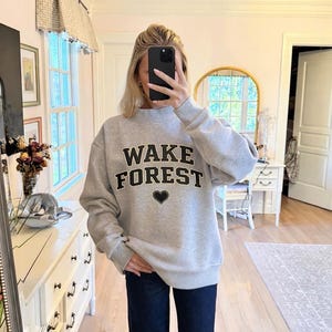 May include: A light gray sweatshirt with the words "WAKE FOREST" in black block letters and a black heart graphic. The sweatshirt has a mock neck and long sleeves.