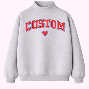 May include: A light gray sweatshirt with a mock neck and long sleeves. The word "CUSTOM" is printed across the chest in red with a blue outline, and a small red heart is centered below the text. A casual, comfortable top.
